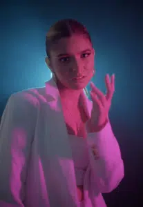 A woman in a white blazer poses under purple and blue lighting, looking at the camera with one hand raised near her face.